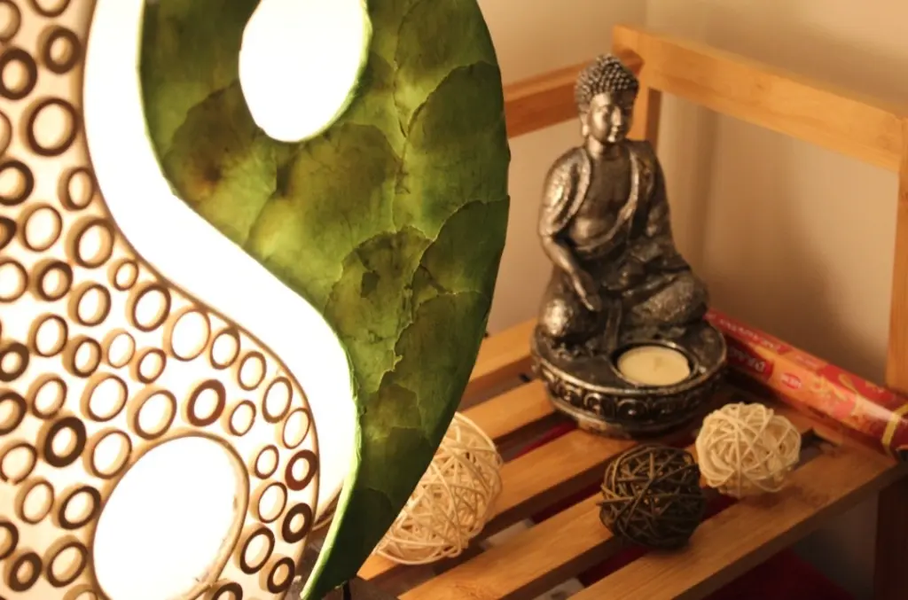 Dieses Bild zeigt einen Buddah mit einem Teelicht, ausstehend im Massageraum der Massageoase im Ergomar Ergolding bei Landshut. Auch eine Ying - Yang Lampe ist zu sehen. Eine Seite der Lampe ist grün gehalten, während die andere Seite mit einem Muster aus Löchern durchzogen ist.
